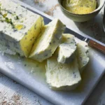 Alt text: Sliced lemon meringue ice cream with lemon balm served on a marble board, garnished with herbs and drizzled with lemon syrup.