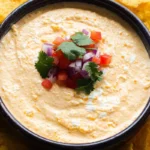 A bowl of creamy cottage cheese queso dip topped with diced tomatoes, red onions, and cilantro, surrounded by crispy tortilla chips.