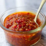 Close-up of a homemade chili garlic sauce recipe in a glass jar with a spoon, showcasing vibrant red color and chunky texture.
