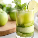 Cucumber Mint Cooler served in a glass with ice, mint sprigs, and lime slice, placed next to green apples on a wooden board.