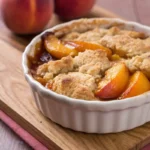 A high-resolution photograph featuring a still-life composition with a rustic and warm aesthetic. The main focus is a bowl containing a dish made from fresh peaches, showcasing a golden-brown, freshly baked crust. The bowl is placed on a wooden surface. The peaches have a rich, warm color, with the peaches and the dish itself being the central elements of the image. fresh peach cobbler