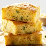 Stack of moist and fluffy slices of Jiffy Mexican cornbread recipe with visible corn kernels and chopped jalapeños on a wooden board.