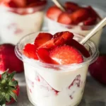 Close-up of a plastic cup filled with fresas con crema recipe, showing layers of strawberries and creamy sweet mixture with a spoon.