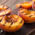 A high-resolution photograph of food items, specifically various types of grilled fruits, primarily peaches, showcasing a golden-brown, well-seared appearance from the grilling process. The fruits are presented on a rustic wooden surface, highlighting the contrast between the natural textures of the wood and the smooth, warm tones of the peaches. The image is detailed, with a focus on the textures and colors of the grilled peaches and the woodgrain patterns. The lighting is warm and inviting, accentuating the rich colors and the appetizing nature of the grilled peaches.