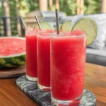 Three tall glasses of watermelon slushie garnished with lime slices, served outdoors on a wooden table—refreshing summer treat from a Watermelon Slushie recipe.