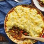 Overhead view of a freshly baked HelloFresh Shepherd’s Pie in a red skillet, with a wooden spoon revealing the hearty beef and veggie filling beneath the mashed potato topping. Perfect for a HelloFresh Shepherd’s Pie Recipe Card.