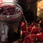 Homemade cherry jam preserves in glass jars surrounded by fresh cherries and a biscuit on a wooden table.