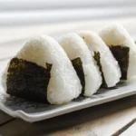 Homemade Japanese Onigiri rice balls wrapped in nori seaweed, neatly arranged on a white ceramic plate.