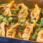Golden baked dumplings in a casserole dish topped with sesame seeds, scallions, and chili oil, a cozy one-pan dumpling bake fresh from the oven.