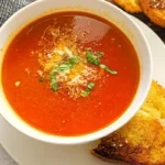 Homemade tomato soup served in a white bowl topped with grated cheese and fresh basil, with toasted bread on the side.