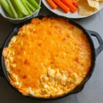 Baked buffalo chicken dip in a cast iron skillet topped with melted cheddar cheese, served with celery, carrots, and tortilla chips on the side.