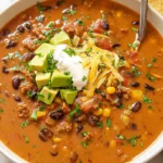 Creamy taco soup in a white bowl topped with diced avocado, shredded cheddar cheese, sour cream, cilantro, and tortilla chips.