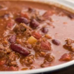 A bowl of thick crockpot chili topped with beans and ground meat, served warm with a spoon