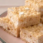Chewy rice krispie treats cut into squares and stacked on a plate, showing their gooey marshmallow texture.