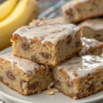 Thick banana bread brownies cut into squares with a soft, moist crumb and glossy chocolate topping, stacked on a plate with visible banana texture and a rich brownie like interior.