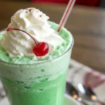 Bright green Shamrock Shake topped with whipped cream and a cherry, served in a glass with a straw, styled with festive St. Patrick’s Day decorations for a fun and vibrant dessert drink.