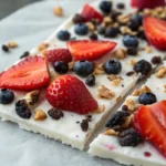 Frozen greek yogurt bark topped with fresh fruits, nuts, and chocolate pieces, broken into chunks and served as a healthy protein dessert snack