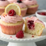 Close-up of a soft pink raspberry buttercream cupcake on a white cake stand, topped with a fresh raspberry and thin lemon wheel , moist raspberry lemon cupcakes recipe with jam-filled center.