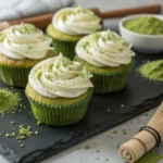 Overhead view of six jade-green matcha cupcakes on a dark slate board, each swirled with ivory white chocolate cream cheese frosting and a dusting of matcha powder, bamboo whisk resting alongside in soft natural light — matcha green tea cupcakes made with a pre-dissolve technique and white chocolate cream cheese frosting from scratch.