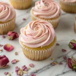 dozen rose vanilla cupcakes on a marble surface, blush pink frosting piped in rose swirls, dried rose petals scattered, natural light, styled beautifully | Alt text: "Rose vanilla cupcakes with pink rosewater buttercream and dried rose petals
