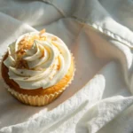 Overhead close-up of a single coconut cream cupcake on white linen, crowned with a swirl of fluffy frosting and a golden toasted coconut top, in warm natural window light — moist coconut cream cupcakes recipe made with a triple coconut method from scratch.