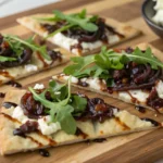 Flatbread on a wooden board, cut into irregular pieces. Dark jammy onions, white ricotta, dark balsamic drizzle, bright green arugula. Shoot slightly angled from above.