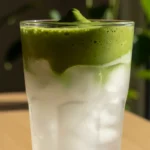 Realistic food photography still life of a sunlit green matcha bubble tea in a tall clear cylindrical glass with clear base and straight sides. The glass contains a bottom two‑thirds water base with six translucent irregular cuboid ice cubes, partially submerged, and a top layer of bright green matcha foam with a smooth slightly mounded undulating surface and small air bubbles. The glass rests on a square tan‑brown kraft‑paper coaster with a coarse fibrous slightly wrinkled texture, which sits on a medium‑brown wooden table showing visible grain, matte finish, curved edge and rounded corners. Behind and to the right of the glass a green plant with multiple dark green elongated leaves is partially visible and out of focus. The background is a smooth matte light off‑white wall. Bright directional sunlight from the left creates sharp angular highlights and diagonal shadows: an unseen object casts a shadow on the wall behind the glass that extends onto the table, and the plant . Coconut matcha cloud