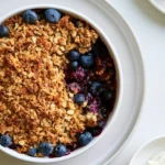 Blueberry crisp with golden oat topping served warm in a white ceramic dish