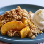 Peach crisp with golden brown oat topping served warm in a white baking dish with a scoop removed