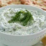 Tzatziki sauce in a white bowl garnished with fresh dill and cucumber slices on a rustic wooden table