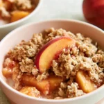 Peach crumble with golden crispy topping served in a white dish with a spoon