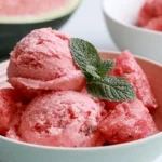 Watermelon ice cream scooped into a white bowl with fresh mint garnish and watermelon slices on a marble surface