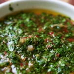 Chimichurri sauce in a white bowl with fresh parsley and grilled steak on a rustic wooden table