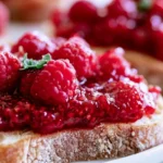 Raspberry jam recipe in a glass jar with fresh raspberries and a spoon on a white marble surface