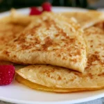 Crepe recipe showing a stack of golden thin French pancakes folded into quarters on a white plate with fresh berries