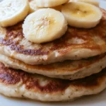 Banana pancakes stacked on a white plate with butter and maple syrup drizzled on top