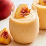 Peach smoothie in a tall glass with fresh peach slices and granola on top