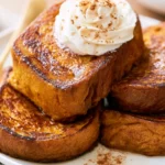 Pumpkin french toast stacked on a white plate with maple syrup drizzle and whipped cream