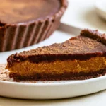 Chocolate pumpkin tart with glossy ganache topping on white plate with gingersnap crust visible