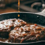 a wide shallow plate or rustic cast iron skillet with two thick, juicy hamburger steaks completely buried under the most gloriously rich, deeply brown caramelized onion and mushroom gravy — a generous pour of extra gravy mid-action for drama