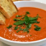 Roasted garlic tomato soup served in a white bowl with fresh basil and cream swirl on top