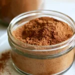Pumpkin pie spice recipe in a small glass jar with a wooden spoon on a rustic table