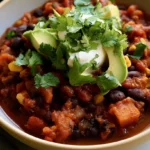 Vegetarian chili in a white bowl with black beans, kidney beans, and diced red peppers visible, topped with sour cream and cilantro