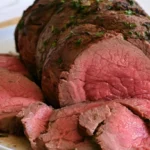 Top round roast sliced on a white platter with fresh rosemary and thyme garnish