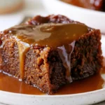 Sticky toffee pudding in a white baking dish with glossy toffee sauce pooled on top and a serving spoon resting in the pan.