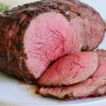 Bottom round roast sliced thin on a white platter with fresh herbs and pan juices