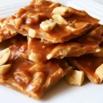 Peanut brittle broken into irregular pieces on a white plate with whole roasted peanuts visible throughout