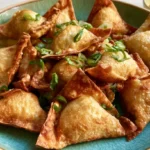 Crab rangoon appetizers on a white plate with golden crispy wonton wrappers and a small bowl of sweet and sour sauce
