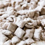 Puppy chow in a white bowl with powdered sugar coating and chocolate visible underneath